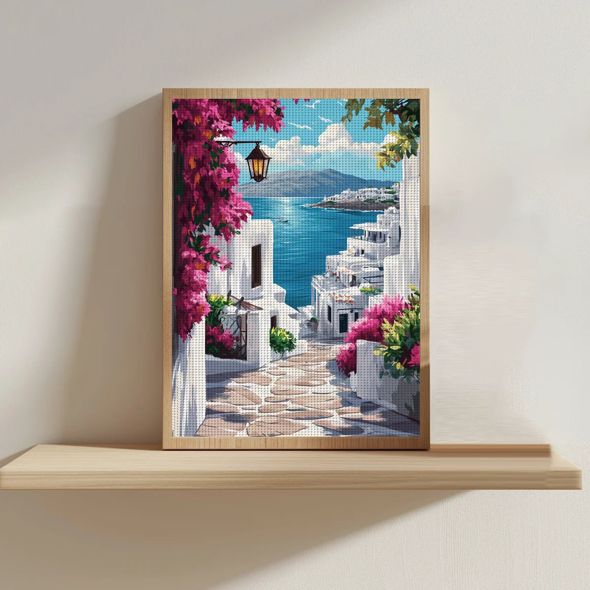 Broderie point de croix paysage de Santorin en Grece encadree et posee sur une etagere en bois flottant contre un mur beige.