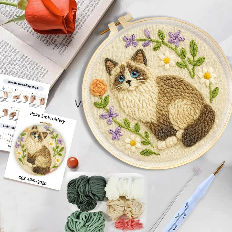 Tambour de broderie punch needle terminé représentant un chat siamois entouré de fleurs, posé sur un bureau à côté de fils de laine.