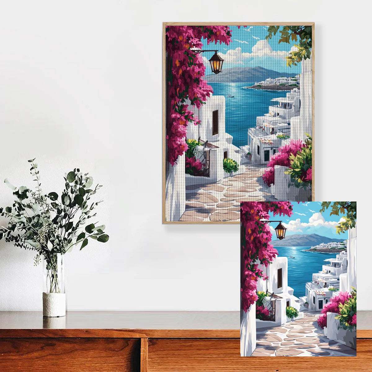 Tableau fait main en point de croix style paysage mediterraneen accroche dans un salon moderne a cote d'un vase et d'une plante.