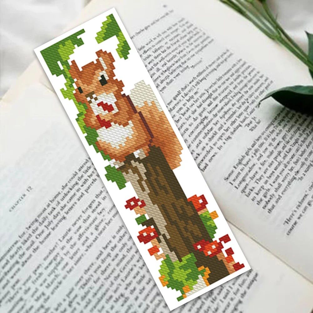 Marque-page brodé en point de croix avec un écureuil mignon sur une souche d'arbre posé ouvert sur un livre.