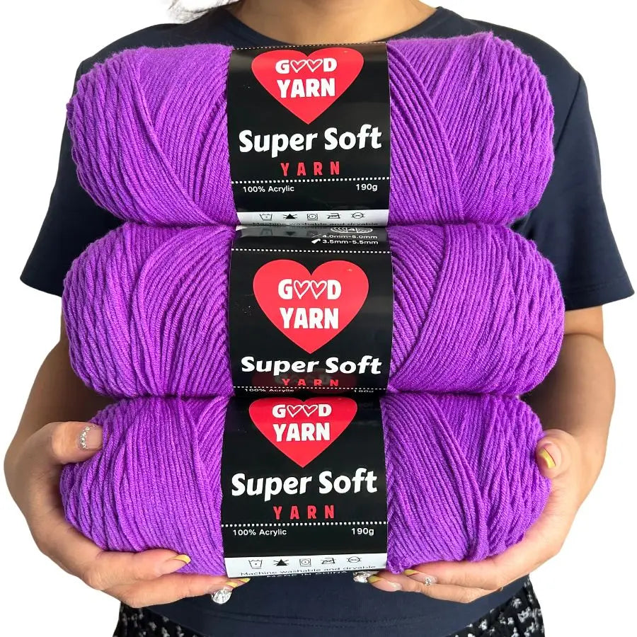 Trois grosses pelotes de laine violettes de la marque Good Yarn Super Soft tenues dans les mains