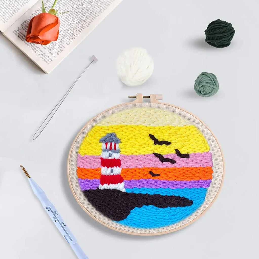 Atelier créatif de punch needle avec un cadre rond illustrant un phare marin au coucher de soleil, entouré de fils et d'outils de couture.