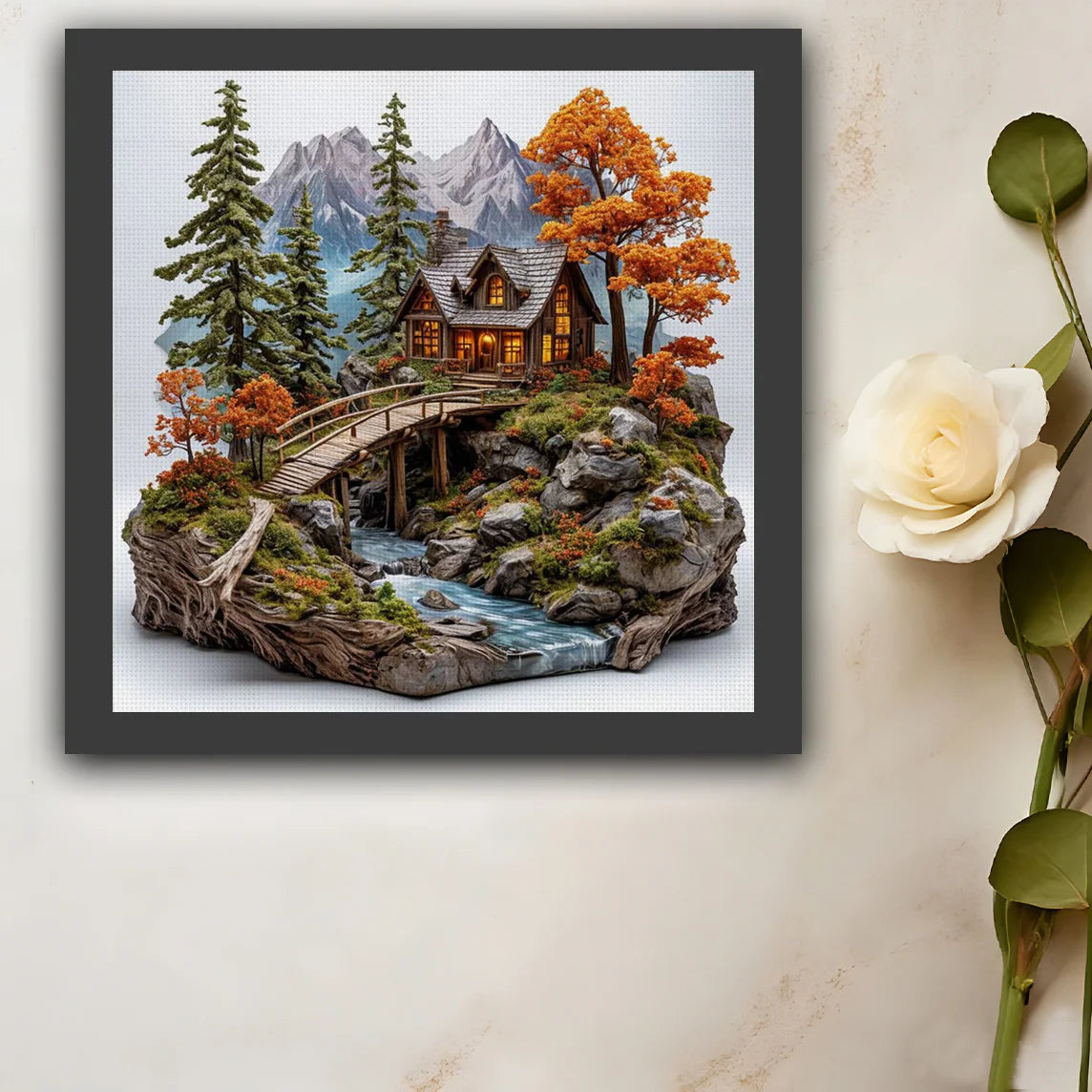 Tableau broderie fait main paysage de chalet alpin encadre en noir suspendu sur un mur a cote d'une rose blanche.