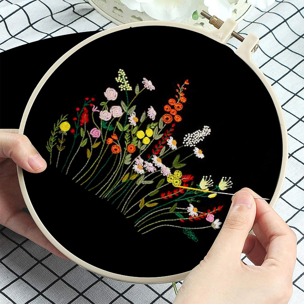 Gros plan sur les mains d'une couturière en train de broder des fleurs des champs colorées au point de tige sur un tissu en toile noire tendue.