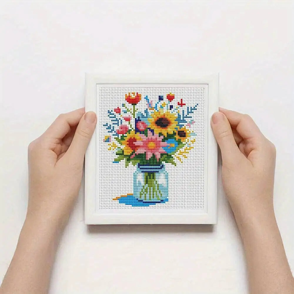 Mains tenant une petite toile de canevas a broder terminee avec un motif de bouquet de fleurs colorees de style champetre.