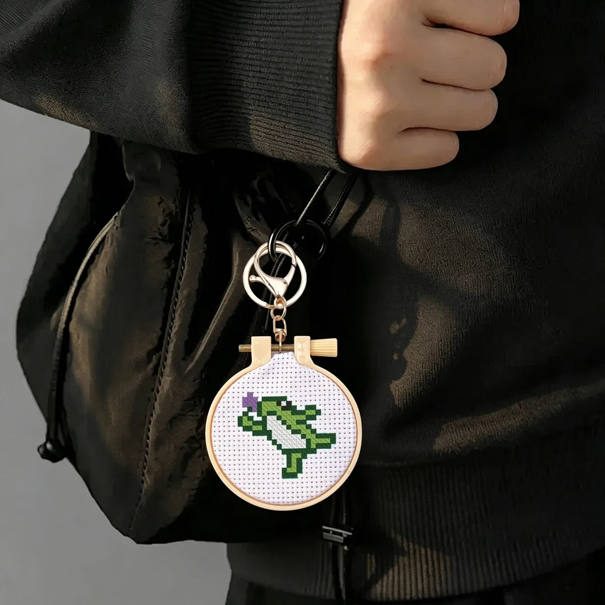 Porte-cle brode en point de croix avec un petit dinosaure rigolo suspendu comme accessoire de mode sur un sac a main noir.